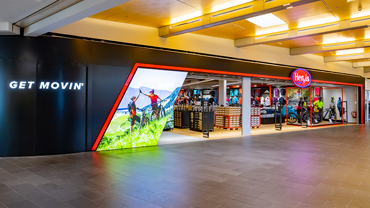 Die Fassade des Hervis Sports Stores in Graz Murpark. Der Store hat ein modernes Design mit einer schwarz-roten Außenansicht. Auf der linken Seite befindet sich ein beleuchtetes Schild mit der Aufschrift 'GET MOVIN''. Der Eingang besteht aus großen Glasfenstern, die verschiedene Sportgeräte und -bekleidung im Inneren des Stores zeigen. Das Innere des Stores ist gut beleuchtet und präsentiert Artikel wie Fahrräder, Kleidung und andere Sportartikel.
