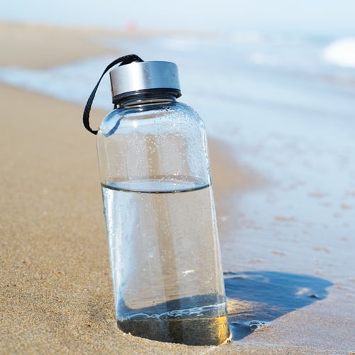 Durchsichtige Wasserflasche mit silbernem Deckel und schwarzem Trageband liegt leicht geneigt im Sand am Strand, mit Wassertropfen auf der Oberfläche und Meereswellen im Hintergrund