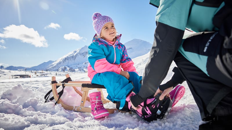 Ein Kind sitzt auf einem Holzschlitten im Schnee, während ihm Eislaufschuhe angezogen werden