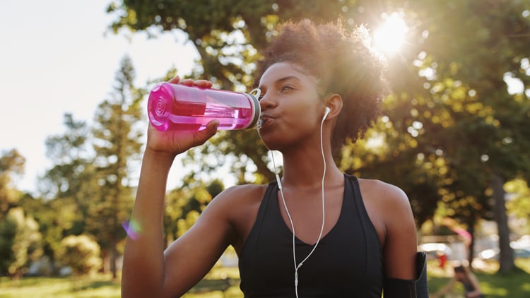 Laufberater Person mit schwarzem Tanktop und Ohrhörern trinkt aus einer pinken Wasserflasche in einem sonnigen Park mit Bäumen im Hintergrund